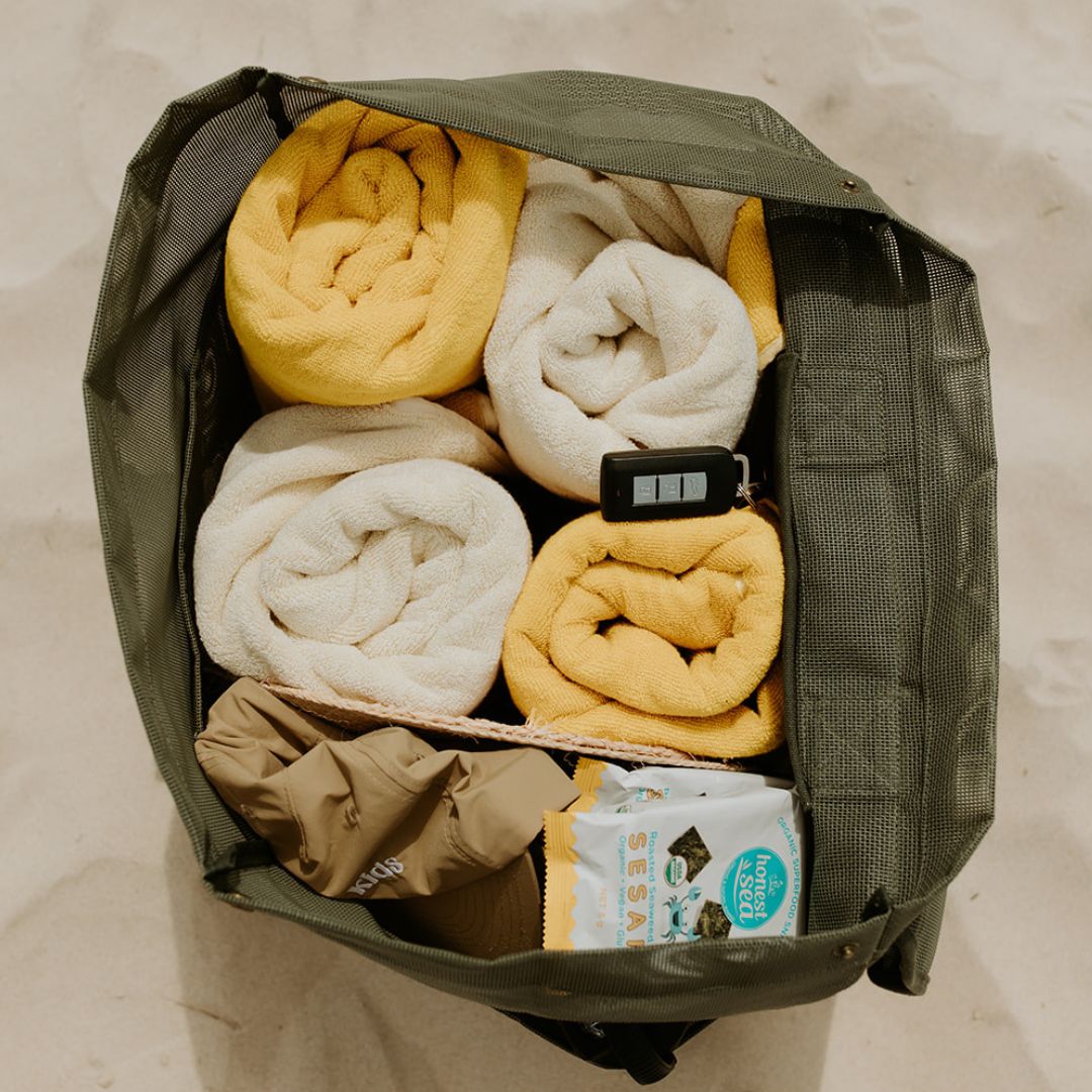 This image shows a top view of the Sande Kids™ Beach Hauler mesh beach bag in dark green. The bag is packed with four bulky beach towels, hats, snacks, and other beach essentials, demonstrating its spacious and functional design.