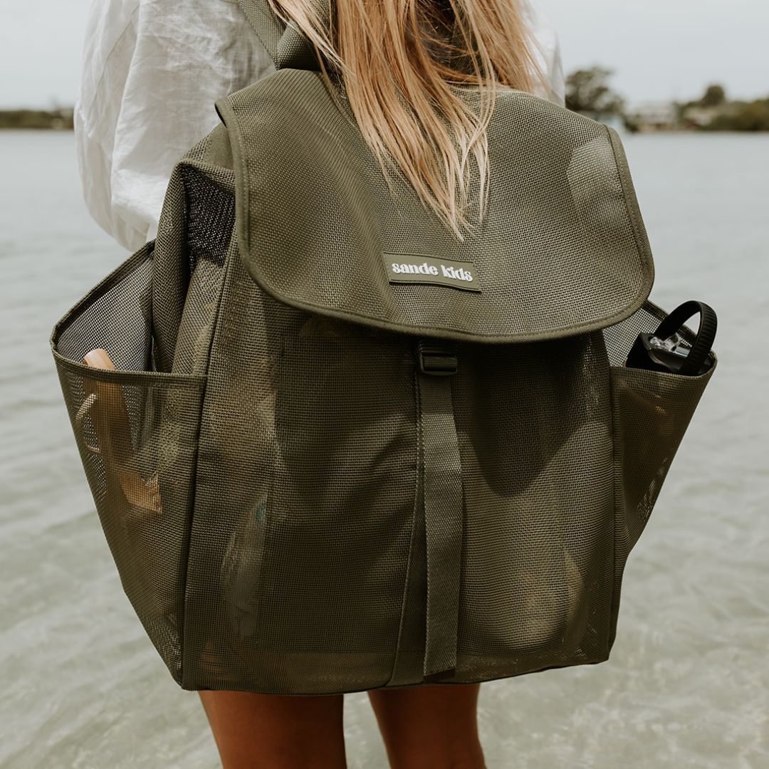 An image of a woman with long blonde hair standing at the water’s edge, wearing the Sande Kids™ Beach Hauler mesh backpack in Pandanus, a dark olive green. The backpack features side pockets, a flap that clips up, and displays a silicone Sande Kids™ logo in white.