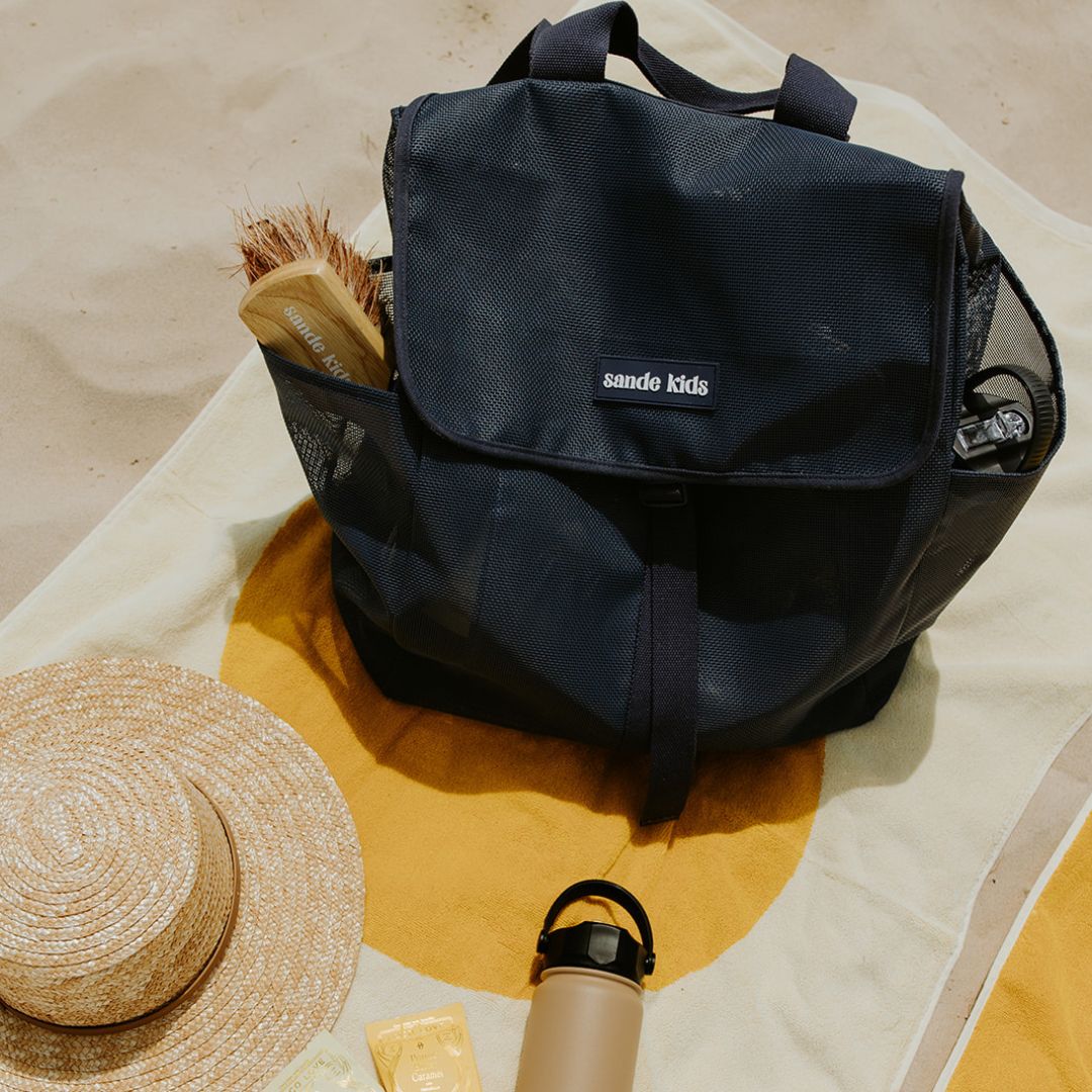 Top view of the Sande Kids™ Beach Hauler mesh backpack, closed and sitting on a beach towel on the sand. A Pargo drink bottle and a Sande Kids™ brush are placed in the outer mesh pockets.