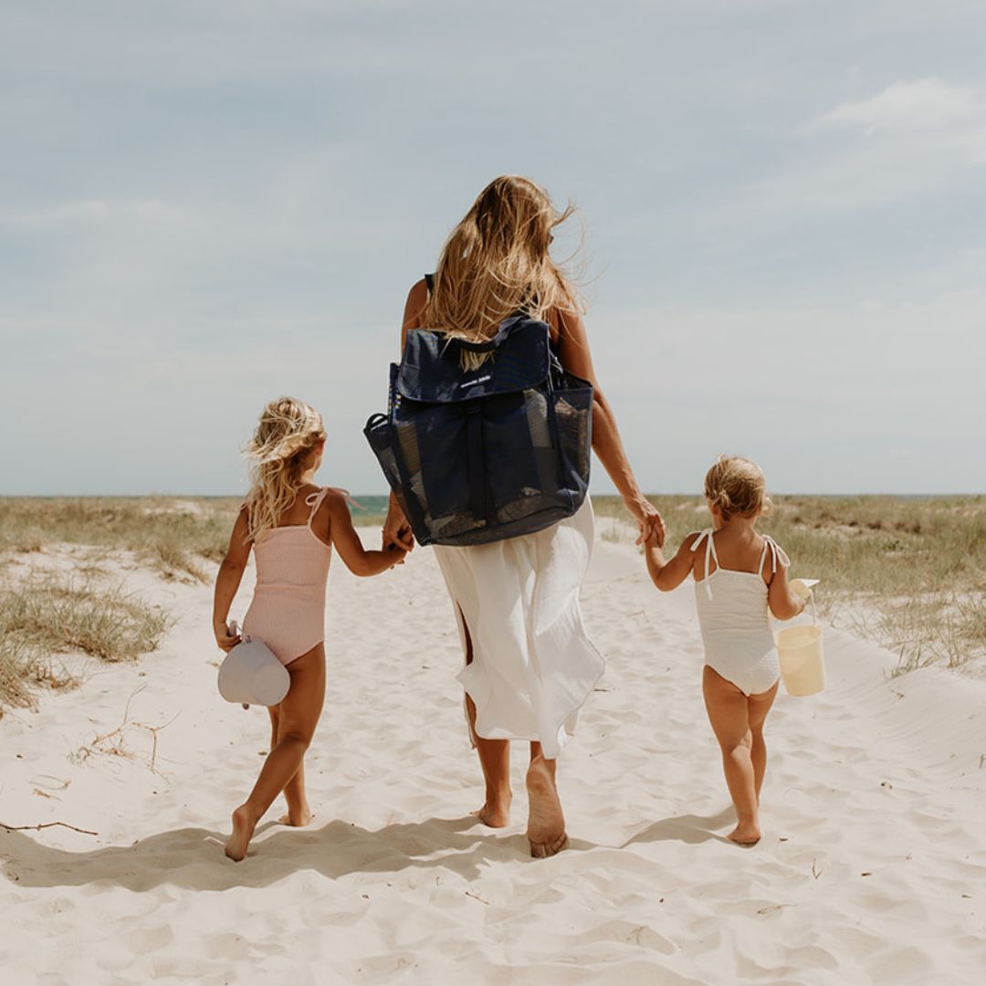 A woman holds the hands of two little girls as they walk along a sandy track to the beach. The mum is wearing the Sande Kids™ Beach Hauler mesh backpack in Deep Sea Navy.