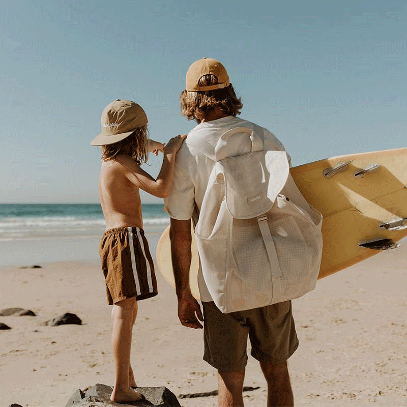 Dad and son checking the surf, with dad carrying a yellow surfboard and the Sande Kids Beach Hauler mesh backpack, and son wearing a Sande Kids merch cap.
