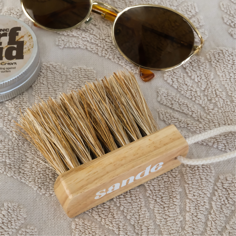 Sande Mini Sand Removal Brush with logo branding, featuring a rubberwood block, coconut and jute fibre bristles, and a cotton cord with brass carabiner, styled on a neutral beach towel with sunglasses and sunscreen.