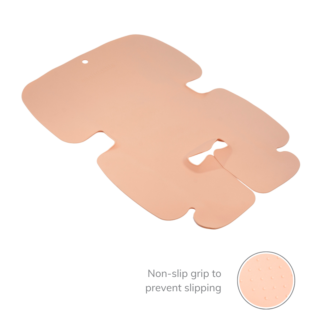 The Sande Kids™ waterproof car seat and pram liner in Coral Pink is shown laid out flat, featuring a molded design around the crotch buckle for added comfort and security. A small inset image highlights the non-stick grip on the back, designed to prevent slipping and ensure a secure fit.