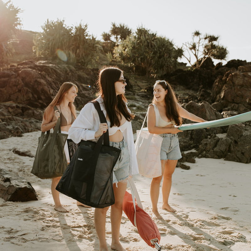 Three friends at the beach, each carrying a Sande Kids Beach Hauler -Mini mesh beach bag in one of our three beautiful colourways: Neutral, Olive Green, and Dark Navy. One girl is carrying a surfboard, while another carries a beach umbrella. They are on the sand with black rocks and pandanus palms in the background.