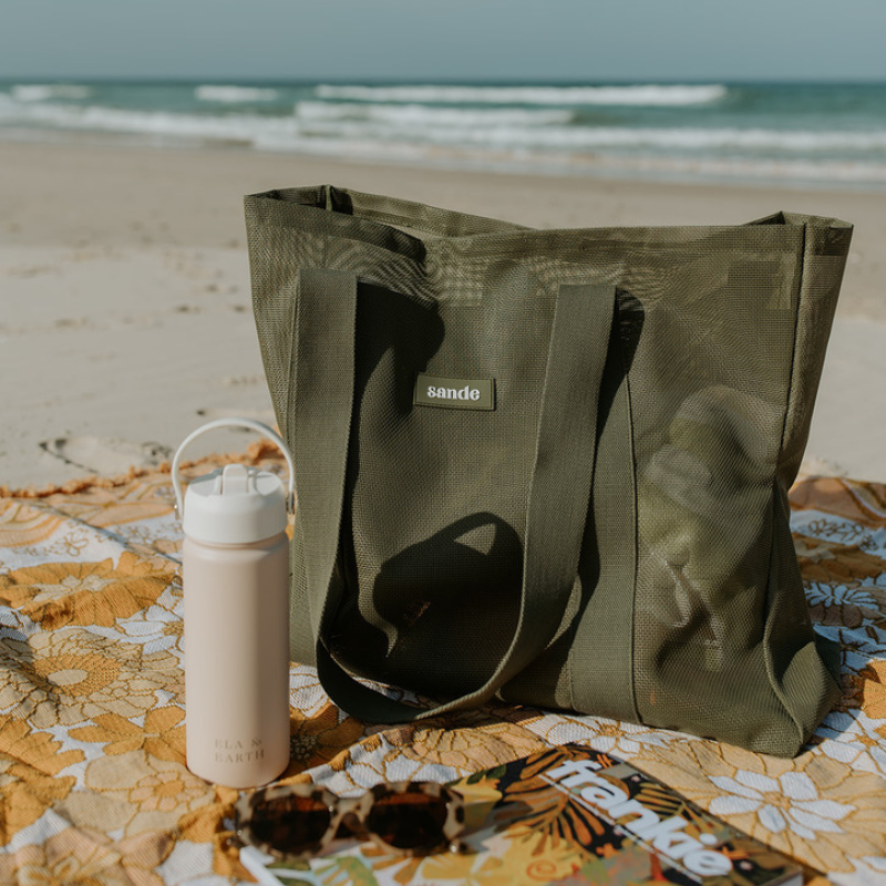 The Sande Kids Beach Hauler - Mini mesh beach bag in Pandanus olive green, sitting on a yellow floral beach blanket at the beach. Ocean in the background.
