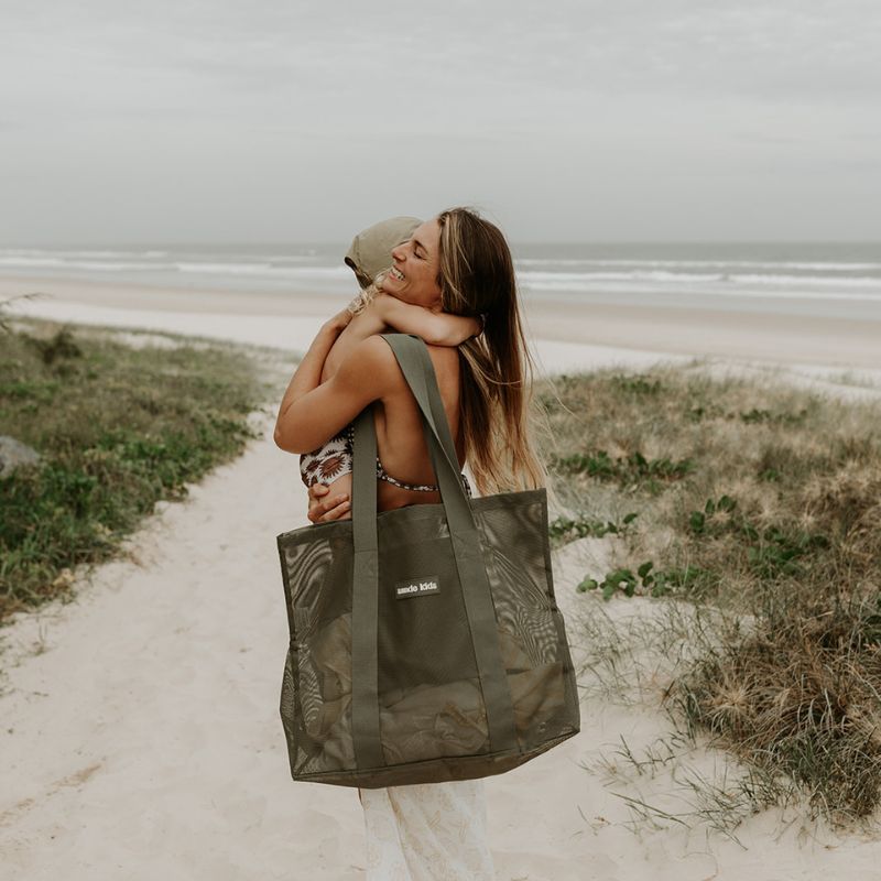 Mother at the beach carrying her toddler son, with a Sande Kids Beach Hauler Mini in Pandanus green on her shoulder, showcasing its convenient size and family-friendly design for beach outings.