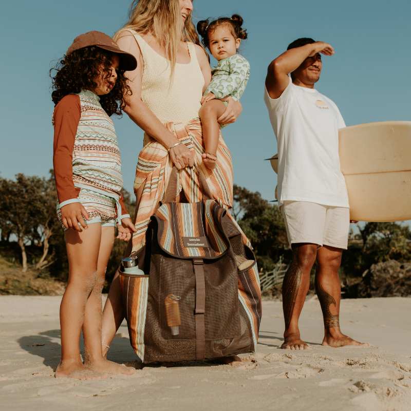 Sande x Banabae mesh beach backpack in brown with retro striped canvas, held by a woman carrying a little girl, while a man holds a surfboard under one arm and a young boy stands beside the woman.