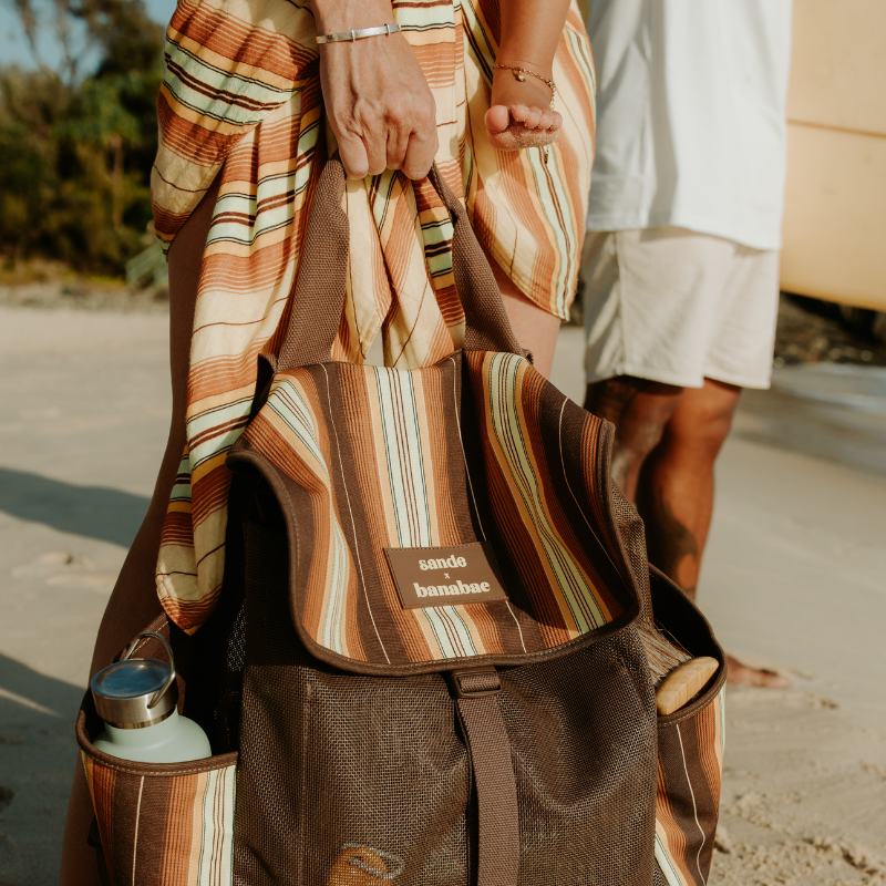 Close-up of a woman holding the Banabae x Sande backpack, with a man carrying a surfboard under one arm beside her.