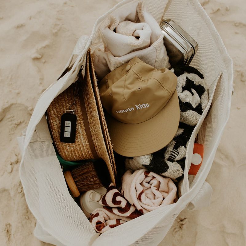 Top view of the Sande Kids Beach Hauler mesh beach bag in Sand, resting on the sand. This image showcases the spacious capacity of this oversized beach bag, highlighting how it easily fits beach towels, sunscreen, hats, snacks, water bottles, and valuables—making it the perfect beach bag for all your essentials.