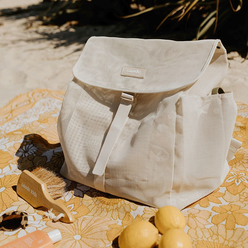 The Sande Kids Beach Mesh Backpack in Sand, sitting on a yellow picnic blanket next to fresh lemons, sunscreen, sunglasses, and the Sande Kids Sand Brush. Vegetation is visible in the background.
