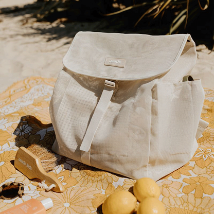 The Sande Kids Beach Mesh Backpack in Sand, sitting on a yellow picnic blanket next to fresh lemons, sunscreen, sunglasses, and the Sande Kids Sand Brush. Vegetation is visible in the background.