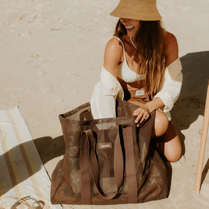 Woman sitting on a sandy beach reaching into an oversized mesh beach bag by Sande in espresso in front of her.