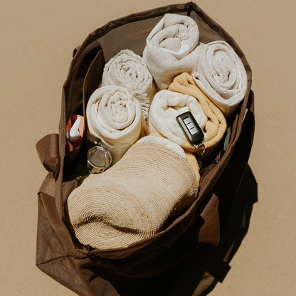 Top view of the Sande oversized mesh beach bag showing capacity with six towels, a sand brush, sunscreen in the internal mesh pocket, and car keys clipped to the key holder. The bag sits on the sand.