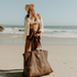 Woman standing on the beach with the ocean in the background, looking up toward the sun. Her hand touches her hair while she holds the Sande oversized Mesh Beach Bag in Espresso, resting on the sand to show its large size.