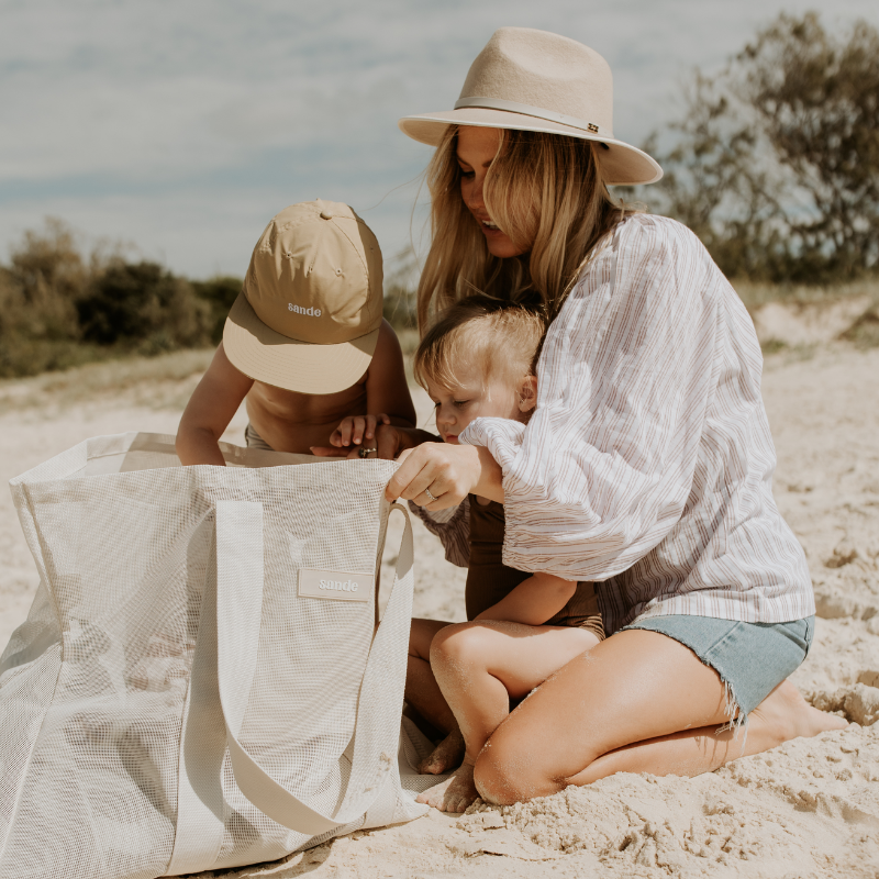 Lifestyle image of a young family looking into their neutral Sande™ mesh beach bag. A little boy wearing a beige Sande™ cap reaches into the bag, while a little girl sits on her mothers lap. The mother has long blonde hair and is wearing a white shirt, denim shorts, and a cream fedora hat.