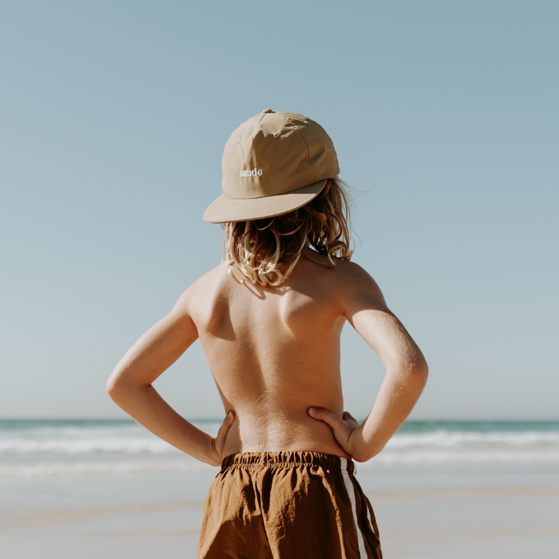 A lifestyle image of a young boy with the beige nylon Sande™ cap worn backwards. He stands on the beach, hands on hip, looking out to the ocean.