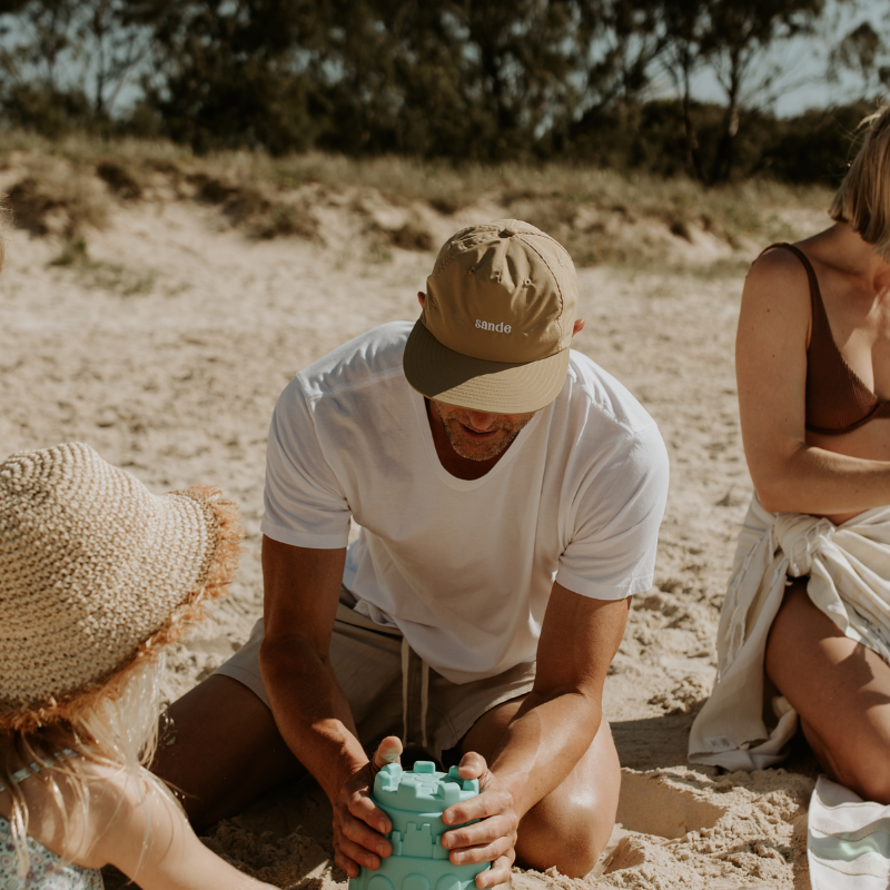 A lifestyle image of dad wearing the beige waterproof Sande™ cap while building sandcastles on the beach with his family.
