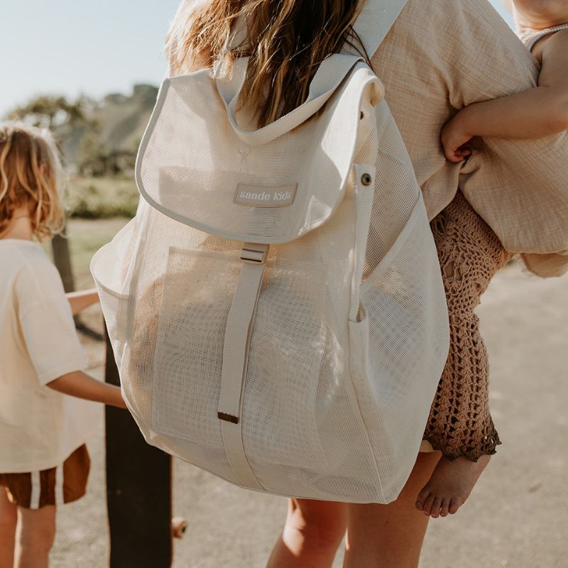 Lifestyle image of mum carrying a toddler on her hip with the Sande Kids Beach Hauler mesh backpack in Sand on her back, ready for a beach day. Features adjustable straps for comfort.