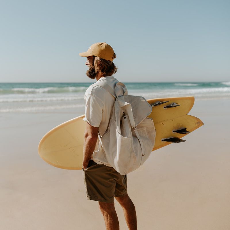 Dad at the beach with Sande Kids Beach Hauler mesh backpack on his back and yellow surfboard under one arm, ready for a beach day.