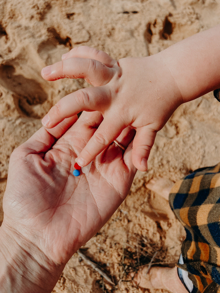 Hands of a parent and child with sand below. Child points to pieces of small plastic rubbish picked up at the beach. Take 3 for the sea.