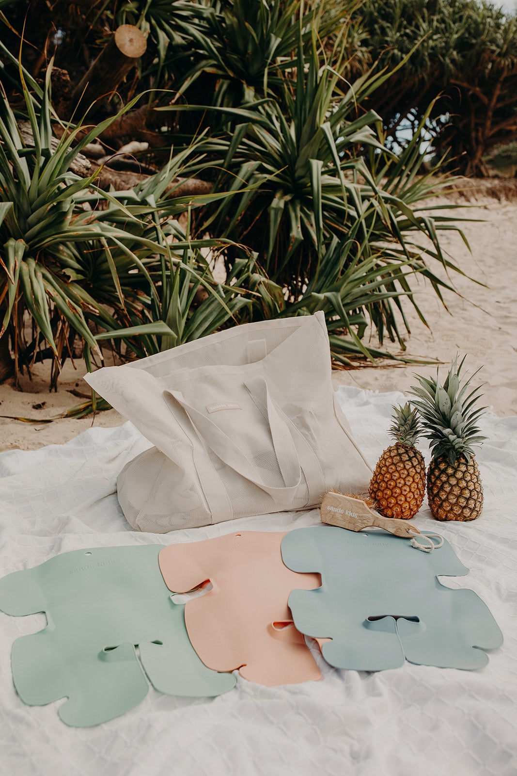 Original Sande Kids product range - Beach Hauler mesh beach bag, sand brush and children's waterproof seat liners. Shown at the beach on a white beach blanket with Pandanus trees in the background.