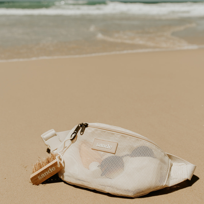 Sande wooden sand removal brush with natural bristles and cotton cord with brass carabiner, clipped to the Sande Essentials Mesh Crossbody in Sand, photographed on sand.

