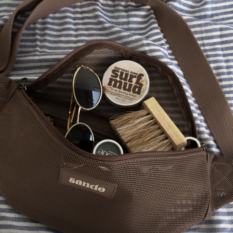Sande Essentials Mesh Crossbody Bag in Espresso, styled on a striped towel with Mini Sand Brush, sunglasses and surf mud sunscreen, all shown inside the crossbody bag.