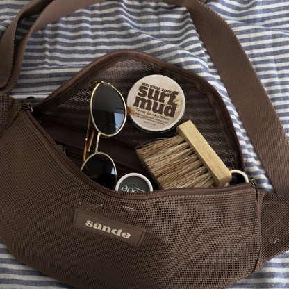 Sande Essentials Mesh Crossbody Bag in Espresso, styled on a striped towel with Mini Sand Brush, sunglasses and surf mud sunscreen, all shown inside the crossbody bag.
