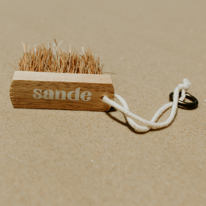 Sande wooden sand removal brush with natural bristles and cotton cord with brass carabiner, photographed on sand.