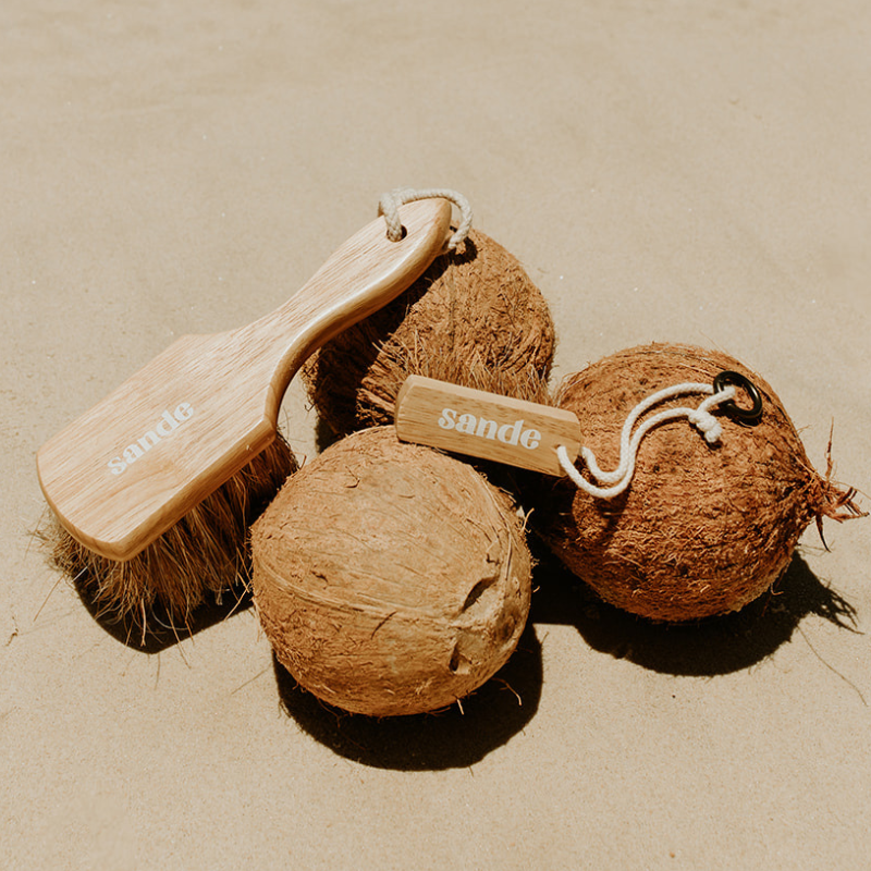 Sande Original Sand Removal Brush and Sande Mini Sand Brush resting on three rough husked coconuts on the sand.