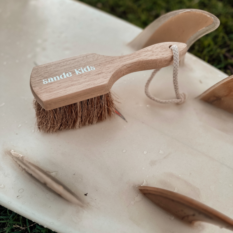 Sande Kids natural sand remover brush sittin on a surf board with natural colour surfboard fins.