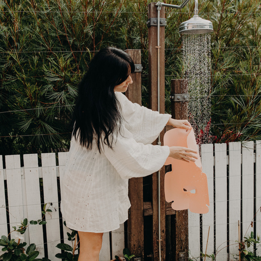 A mother rinsing their Sande Kids™ Waterproof Car Seat and Pram Liner under a garden shower after returning from the beach with her toddler.