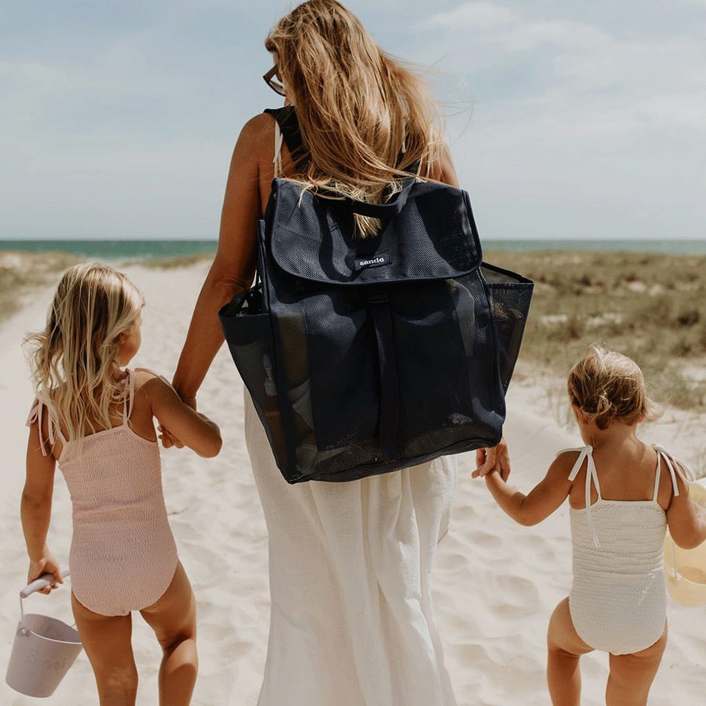 A woman holds the hands of two little girls as they walk along a sandy track to the beach. The mum is wearing the Sande Kids™ Beach Hauler mesh backpack in Deep Sea Navy.