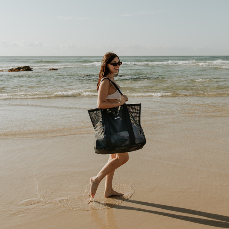 A young woman carrying the Sande Kids Beach Hauler - Mini mesh beach bag in Deep Sea Navy along the shoreline at the beach. The transparent mesh reveals beach towels and other essentials inside the bag.