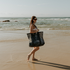 A young woman carrying the Sande Kids Beach Hauler - Mini mesh beach bag in Deep Sea Navy along the shoreline at the beach. The transparent mesh reveals beach towels and other essentials inside the bag.