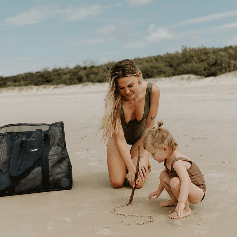 A mum and little girl at the beach, drawing with a stick in the sand. Next to them is their Sande Beach Hauler mesh beach bag in Deep Sea Navy, resting on the sand.