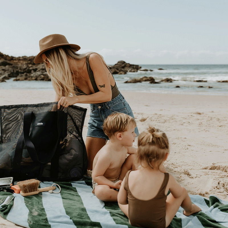 A mum and two little kids at the beach, sitting on a green and blue striped beach towel. The mum is rummaging through her oversized mesh beach bag by Sande Kids, the Beach Hauler mesh bag in Deep Sea Navy. The ocean is visible in the background.