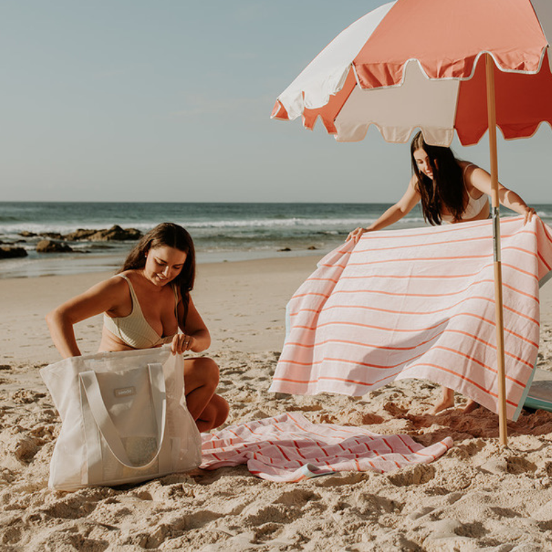 Friends enjoying a day at the beach with their neutral Sande Kids Mini beach bags. The scene features pink and red striped beach towels laid out alongside a coral red and white beach umbrella, creating a vibrant and fun atmosphere for a perfect beach day.