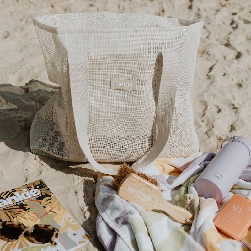 Sande Kids mesh beach bag in neutral colour Sand Dune displayed at the beach, ideal for a solo beach trip. The bag is packed with essentials including a magazine, sunscreen, water bottle, towel, and Sande Kids sand brush, showcasing its practicality and style for beach outings.