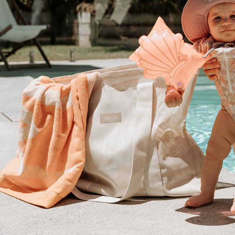 Baby girl at the pool wearing pink shell floaty bands, with the Sande Kids Beach Hauler Mini mesh beach bag in Sand colorway nearby. This image highlights the perfect combination of fun and practicality for family pool days, featuring the stylish and durable beach bag.