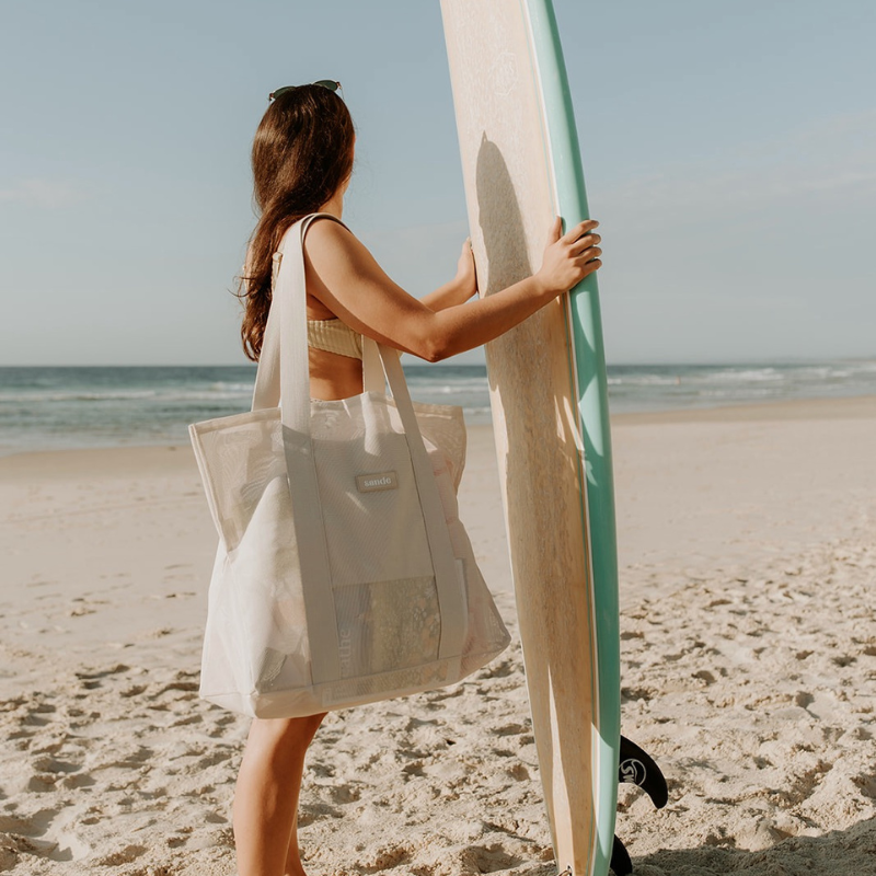 Sande Kids Beach Hauler Mini in Sand colorway, carried by a woman holding her turquoise and timber longboard upright while checking the surf. This image showcases the stylish and functional mesh beach bag, perfect for beachgoers looking for convenience and style during their outings.