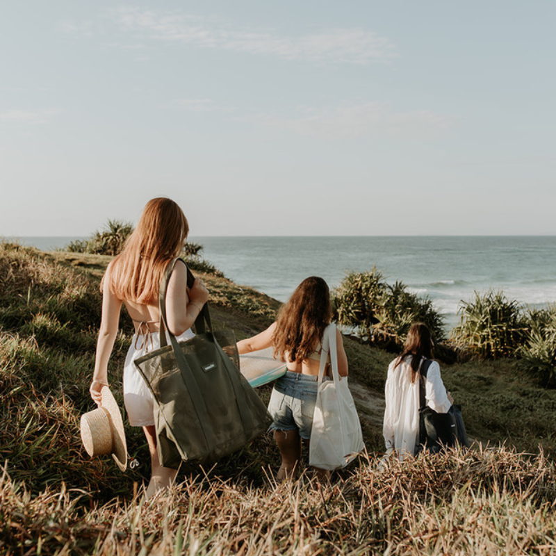 A group of girlfriends walking down a grassy headland at Cabarita Beach, carrying surfboards and their Sande Kids Mini beach bags. This scene captures the essence of fun and friendship at the beach, showcasing the stylish and functional design of the Sande Kids bags.