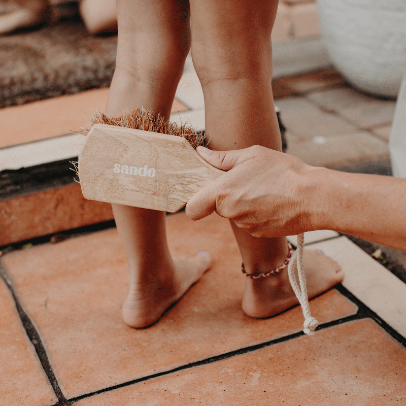 A lifestyle image of a little girl standing on terracotta stairs at the front door, with her legs being swept of sand. The Sande™ Sand Brush is used to gently remove the sand after a day at the beach.
