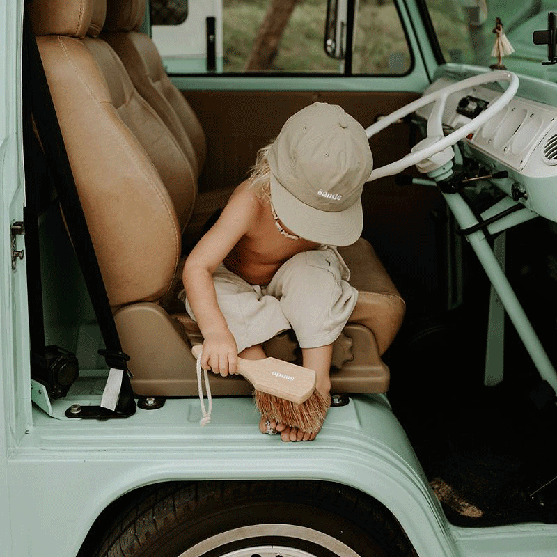 Little boy siting at the door of a vintage Kombi Van using the Sande Kids sand brush to sweep sand from his feet.