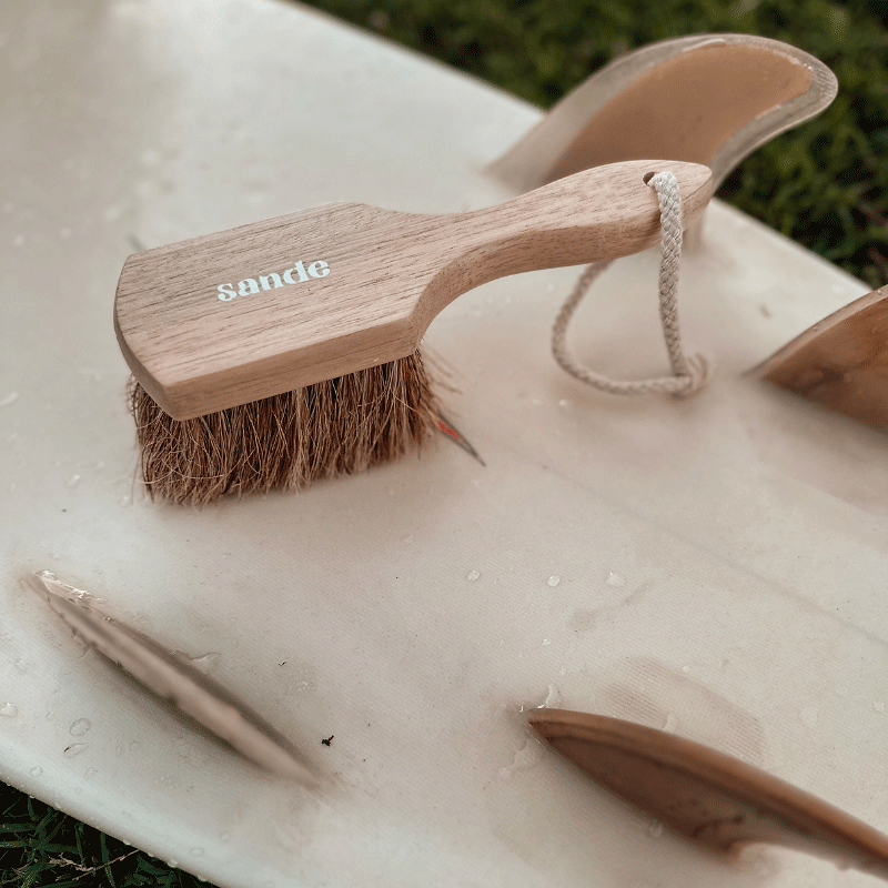 Sande natural sand remover brush sittin on a surf board with natural colour surfboard fins.