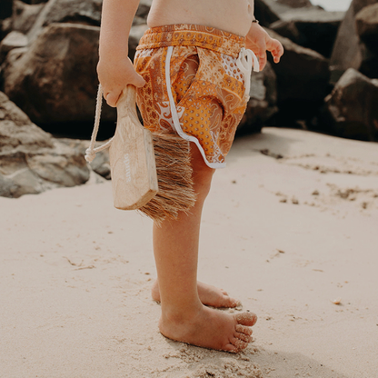 A little boy standing on the beach, holding the Sande™ Sand Brush. Wearing orange board shorts, he’s ready to sweep sand off his feet, with black rocks in the background.