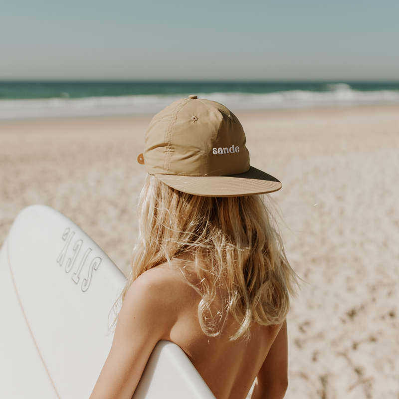 Lifestyle image of a young surfer with long blonde hair, holding a surfboard under one arm on the beach, looking at the ocean, wearing a beige quick-dry Sande™ cap backwards.